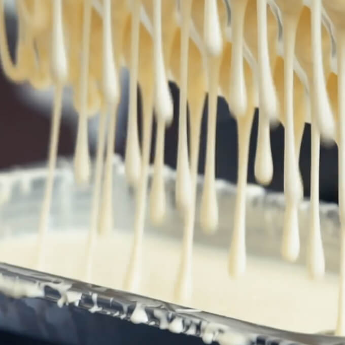 Close-up of dripping creamy white sauce or batter from a perforated utensil into a container.