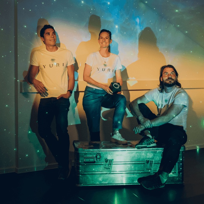 Three people wearing white Yuri t-shirts posing indoors against a wall with star-like light projections, one sitting on a metal box holding a device.